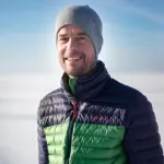 Person med lue og vattert jakke smiler mot kameraet i et snødekt landskap under klar himmel.