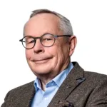 Person med briller og kort hår smiler mot kameraet, iført blazer og skjorte foran en lys bakgrunn.