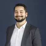 Smilende person i dressjakke og skjorte står foran en blå bakgrunn.