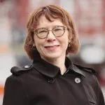 Portrett av en kvinne med kort, brunt hår og briller, iført mørk jakke, som smiler utendørs med bymiljø i bakgrunnen.