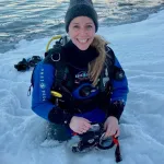 Nina Jensen i dykkerdrakt, sittende i snøen ved sjøen under et dykk, smilende mot kamera.
