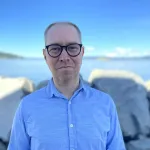 Portrett av en mann med briller i lyseblå skjorte ved sjøen.