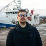 En person med kort hår og briller står utendørs på en kai. I bakgrunnen ligger et stort skip fortøyd ved havn, med kran og overbygg synlig. Personen har på seg en mørk jakke, og det er overskyet vær.