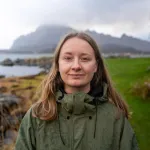 Kvinne med langt, lysebrunt hår og grønn jakke står ved en steinete kyst. I bakgrunnen ses hav, grønne områder og fjell under en overskyet himmel.