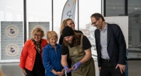 En kokk viser frem mat til en gruppe smilende personer under et arrangement, med bannere i bakgrunnen.