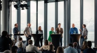 Syv paneldeltakere står samlet bak høye bord på en scene under en konferanse, med et publikum foran seg. Store vinduer i bakgrunnen viser snødekte fjell og hav. En deltaker til høyre snakker mens de andre lytter.