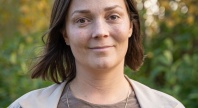 Portrett av en kvinne utendørs, med kort brunt hår, beige jakke og brun topp, foran grønn og gyllen høstlig bakgrunn.
