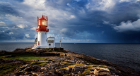 Fyrtårn med hvit base og rød topp står på en klippe ved havet, med dramatisk himmel og skyer i bakgrunnen.