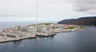 Oversiktsbilde av et stort industrikompleks ved kysten, med røyksøyle fra en pipe og fjell i bakgrunnen.
