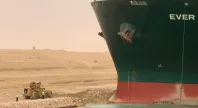 Et stort containerskip sitter fast på grunn i en kanal, mens en gravemaskin og arbeidere står ved siden av for å prøve å frigjøre det.