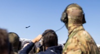 oldater med hørselvern og kamuflasjeuniform ser på et jagerfly som flyr over et fjellområde under en militær øvelse.