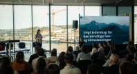En kvinne står og holder innlegg foran et publikum i et lyst lokale med store vinduer mot havna i Tromsø. På en stor skjerm ved siden av henne står teksten «Ungt lederskap for bærekraftige hav- og klimaløsninger», med logoen til Senter for hav og Arktis. Publikum følger med på presentasjonen, og bakgrunnen viser bygg, båter og en kran ved havna.