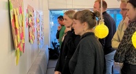 En gruppe deltakere står samlet foran en vegg med store ark dekket av fargerike post-it-lapper, og diskuterer ideer i en workshop-setting. Stemningen er konsentrert og engasjert