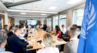 Møte i konferanserom med mange deltakere samlet rundt et langt bord, presentasjon vises på skjerm i enden av rommet, med en FN-flagg synlig i forgrunnen.