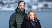 To personer står utendørs foran sjø og snødekte fjell, en mann i grønn dunjakke og briller og en kvinne i mørk kåpe og skjerf.