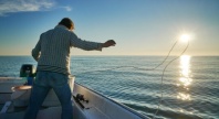 En person står i en liten båt og kaster en line ut i havet mens solen står lavt over horisonten. Sollyset glitrer i det rolige vannet og skaper en varm stemning i kveldssolen.