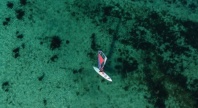 Luftfoto av en person som seiler på et brett med fargerikt seil over grunt, turkist vann. Havbunnen med mørke flekker av tang og stein skimtes tydelig gjennom det klare vannet.