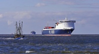 Et stort lasteskip med teksten "ML RORO" på skroget seiler på havet, mens en mindre fiskebåt passerer i forgrunnen. I horisonten skimtes flere fartøy under en delvis skyet himmel.