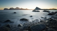 Kystlandskap i solnedgang med steiner i forgrunnen og fjell i horisonten. Lang eksponering gir havet et sløret, tåkete uttrykk som kontrasterer mot de mørke fjellformasjonene i bakgrunnen.