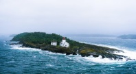 En liten øy med et hvitt fyrtårn og bolighus omgitt av grønn vegetasjon, ligger ute i et opprørt hav. Bølgene slår mot klippene, og tåke og gråvær gir landskapet et dramatisk kystpreg.