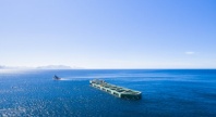 En oppdrettsmerd slepes utover havet av en båt under klar blå himmel. Det dype blå vannet strekker seg mot horisonten, der fjellene skimtes i det fjerne.