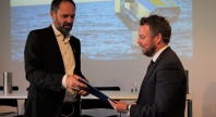 To menn i dress står på en scene under en presentasjon, der den ene overrekker et dokument til den andre. I bakgrunnen vises et bilde av havet og en kikkert på storskjerm.