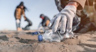 Hånd med hanske plukker opp en plastflaske fra sanden under strandrydding, med flere personer som rydder i bakgrunnen.