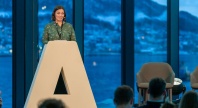 En person står på en scene bak en stor talerstol formet som en “A”, og holder et innlegg foran et publikum, med vinterlandskap og fjell synlig gjennom store vinduer i bakgrunnen.