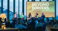Et panel med fem personer sitter på en scene foran et publikum under Arctic Frontiers 2025, med et stort bakskjermbanner som viser teksten “Beyond Borders” og utsikt over fjell og hav gjennom de store vinduene i bakgrunnen.