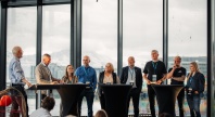 Et panel med ni personer fra ulike deler av sjømatnæringen og forvaltningen står samlet foran store vinduer med utsikt mot fjell og by. De deltar i en panelsamtale foran publikum under en konferanse.