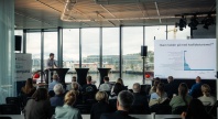 En foredragsholder presenterer for et publikum under et Blått Kompass-arrangement, med en stor skjerm som viser statistikk om havfisketurisme og utsikt mot havn og snødekte fjell gjennom vinduene bak.
