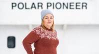 En kvinne med grå strikkelue og brun ullgenser med hvitt mønster står ute foran et skip med navnet «POLAR PIONEER» i bakgrunnen.