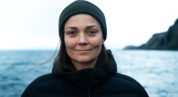 Person iført mørk jakke og grønn lue står utendørs foran et hav og fjell i bakgrunnen på en overskyet dag.