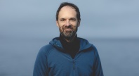 En mann står utendørs foran havet og smiler mot kameraet. Han har på seg en blå hettegenser og har kort hår og skjegg.