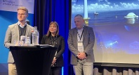 Tre personer står på en scene under et arrangement ved UiT Norges arktiske universitet; en presentasjons­skjerm bak dem viser et havmiljø med offshore vindmølle og havinstallasjoner.