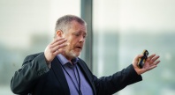 Mann med skjegg og mikrofon gestikulerer engasjert mens han holder en presentasjonsklikker.