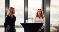 To unge kvinner står på en scene bak et høyt rundt bord under en paneldiskusjon. Begge har mikrofoner og ser konsentrerte ut. I bakgrunnen sees store vinduer med utsikt over snødekte fjell og fjord.