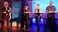 Fire personer deltar i en paneldebatt på en scene under arrangementet «Kystpuls», arrangert av Senter for hav og Arktis. En mann står med mikrofon til venstre, mens tre personer står bak høye bord foran en skjerm med arrangementets logo.