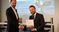 To menn i dress håndhilser mens den ene holder en rapport med tittelen «Sustainable Blue Economy in the Norwegian Arctic». Et bilde av havet vises på skjermen bak dem.