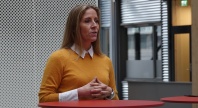 Kvinne i sennepsgul genser står bak et rødt bord og snakker innendørs, med perforert vegg og kontormiljø i bakgrunnen.