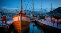 Trebåt fortøyd ved kai i vinterlys i Tromsø havn, med Tromsøbrua og snødekte fjell i bakgrunnen.