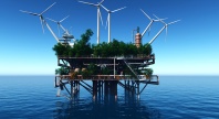 En ombygd, rusten havplattform ute på åpent hav, dekket av trær og vegetasjon, med flere store vindturbiner montert rundt strukturen under en klar blå himmel.