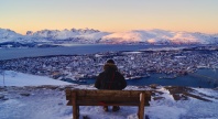 En person sitter på en benk og ser ut over Tromsø by, omgitt av snødekte fjell og hav i solnedgang.