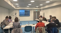 Mennesker samlet rundt bord formet som hestesko. En skjerm i front. 