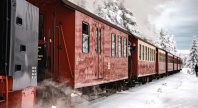 Et damptog med røde og gule vogner kjører gjennom et snødekt landskap omgitt av trær. Dampen stiger fra toget mens en person tar bilder ut av vinduet.