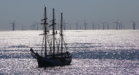Et seilskip ligger på glitrende hav med en rekke vindmøller til havs i bakgrunnen, som illustrerer kontrasten og sameksistensen mellom tradisjonell sjøfart og moderne fornybar energi.