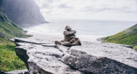 En liten varde av steiner står på en fjellhylle med utsikt mot havet og en strand omgitt av grønne fjellsider.
