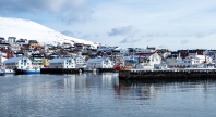Kystby med fargerike hus og fiskebåter ved kai, omgitt av snødekte fjell og rolig hav i nordnorsk vinterlandskap.