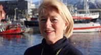 Kvinne med blondt hår smiler foran en havn med båter og bygninger i bakgrunnen på en solrik dag.