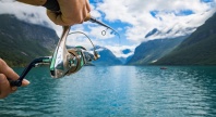 En person fisker med en snell i klart vann omgitt av fjell og solskinn, trolig i en norsk fjord.